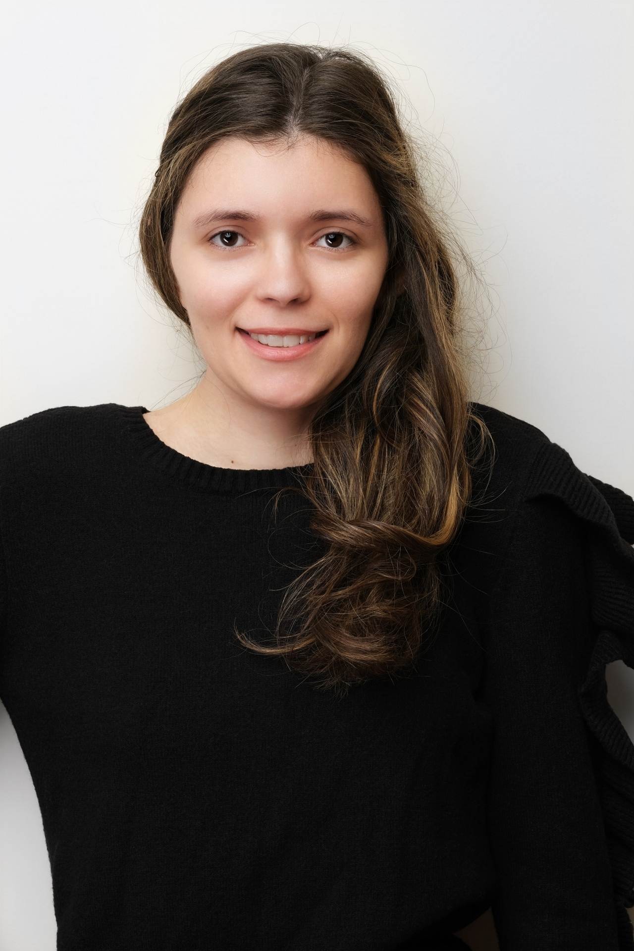 A confident woman with a bright smile poses in a black sweater and pink lipstick against a smooth, light grey background.